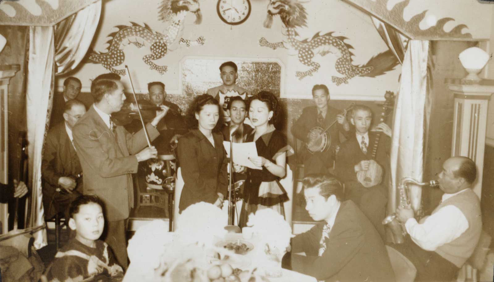 <span style="font-weight: 400;">Photo sépia montrant l’orchestre disposé en demi-cercle autour de deux chanteuses, tandis que Raymond Jai joue du violon à leurs côtés.</span>