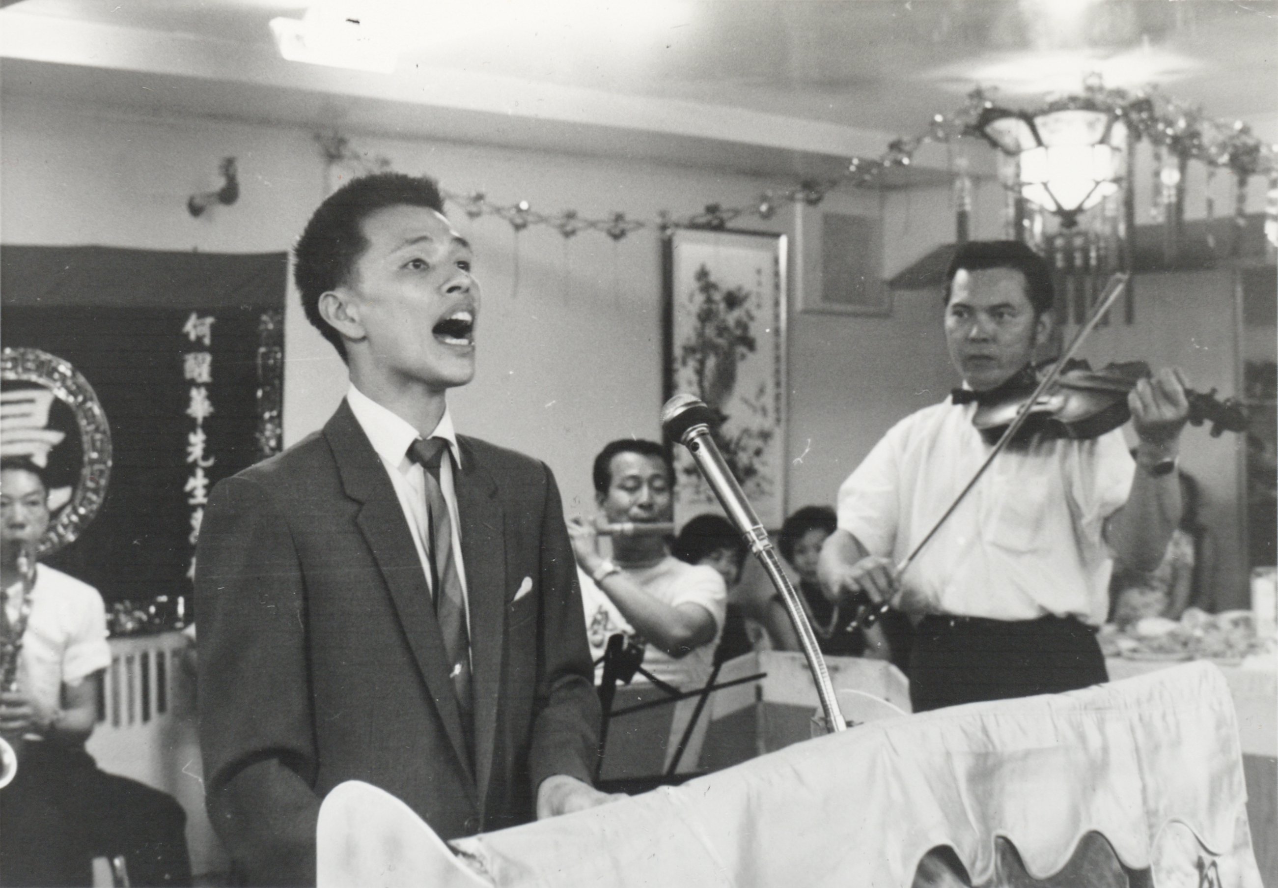 <span style="font-weight: 400;">Un jeune homme en costume chante aux côtés d’un violoniste et d’un petit orchestre.</span>