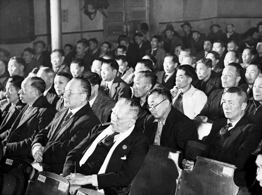<span style="font-weight: 400;">A black-and-white photograph of a full house at the Sing Kew Theatre. The audience is predominantly men dressed in western-style suits, looking engaged.</span>