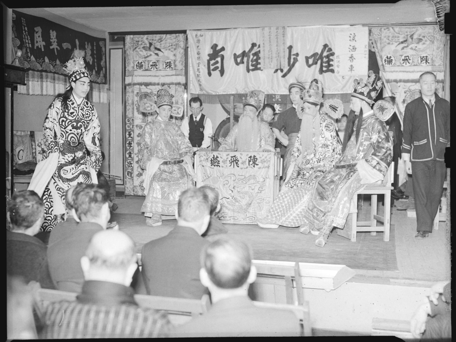 <span style="font-weight: 400;">Performers in embroidered robes and ornate headdresses pose on an elevated wooden stage, resembling a historical courtroom drama. Some depict figures with long white beards and formal court costumes, while colourful painted backdrops and hanging banners add to the scene. Audience members in Western suits closely observe from the foreground.</span>