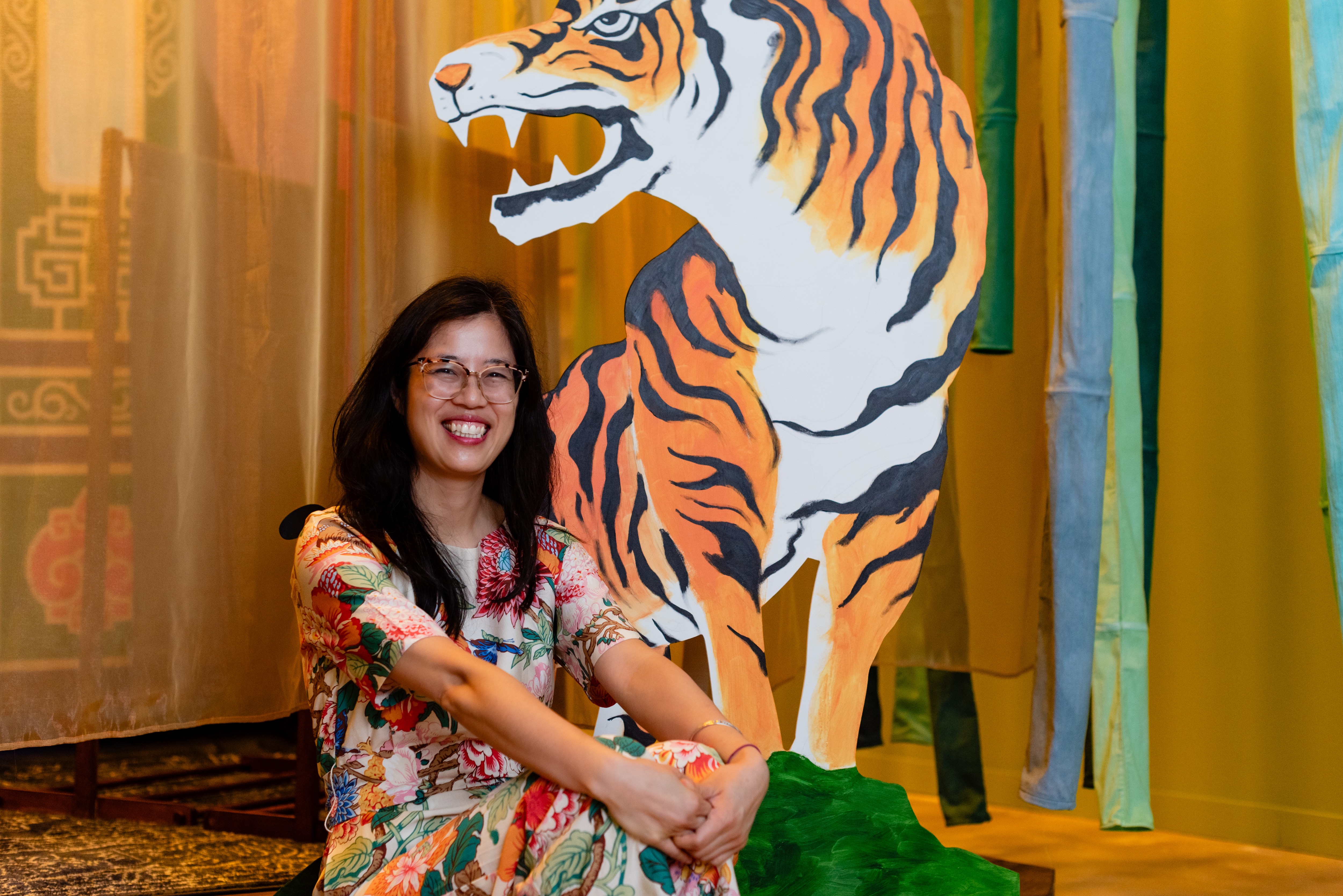 <span style="font-weight: 400;">Karen est assise sur scène, regardant l’appareil photo, vêtue d’une robe colorée à manches courtes et portant des lunettes. Derrière elle se trouve une grande silhouette de tigre peinte à la main, créant un arrière-plan attrayant.</span>