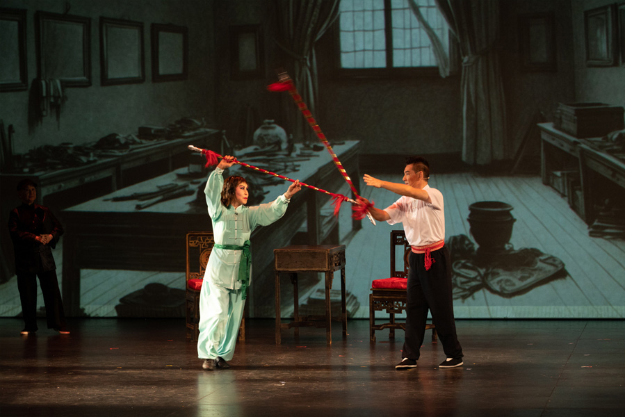 <span style="font-weight: 400;">Deux artistes s’affrontent avec des lances rouge et blanc. Un décor d’intérieur avec des tables et des accessoires est projeté en arrière-plan.</span>
