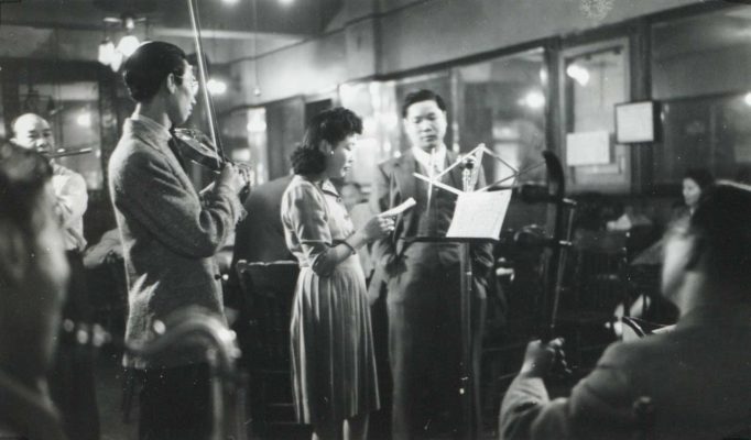 <span style="font-weight: 400;">Photo en noir et blanc au flou artistique d’une femme vêtue d’une robe de style occidental des années 1930, aux cheveux permanentés, debout devant un pupitre tandis que des personnes jouent de la musique autour d’elle.</span>