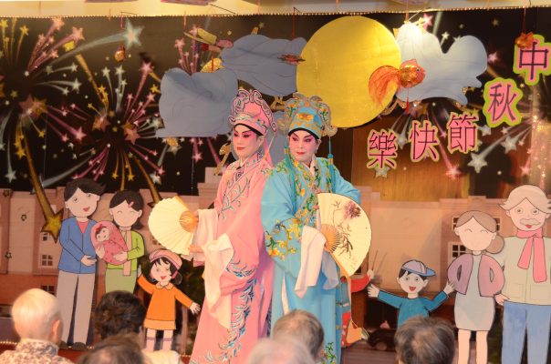 <span style="font-weight: 400;">Deux artistes portant des costumes assortis rose et bleu posent avec des éventails et des manches d’eau flottantes. À l’arrière, la scène est décorée de grandes silhouettes de personnages de style </span><i><span style="font-weight: 400;">anime</span></i><span style="font-weight: 400;"> (animation japonaise), de feux d’artifice et de caractères chinois.</span>