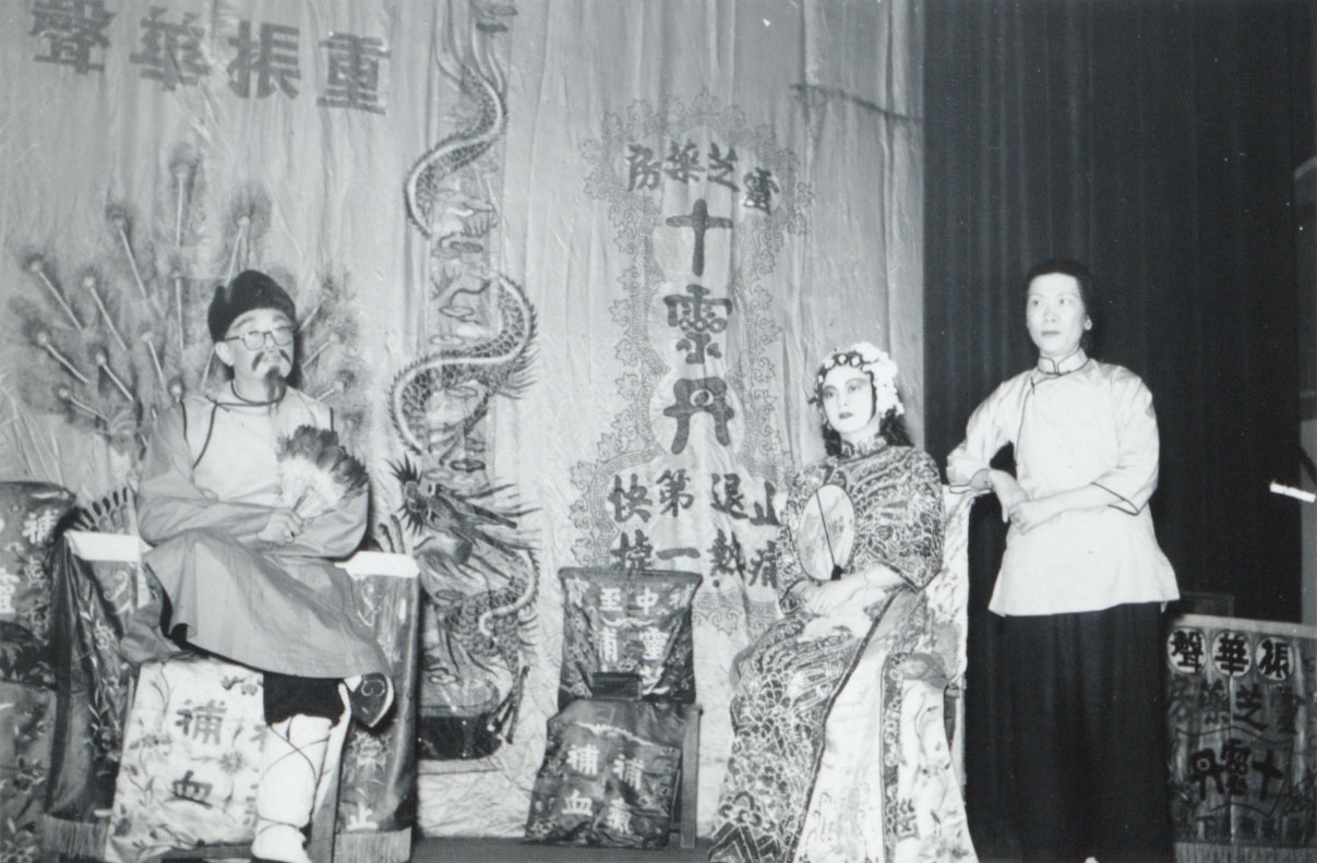 <span style="font-weight: 400;">Trois artistes sont sur scène, incarnant respectivement un rôle masculin portant un chapeau et un costume simples, un rôle féminin dans un costume richement décoré, et une femme, également vêtue d’un costume sobre. </span>