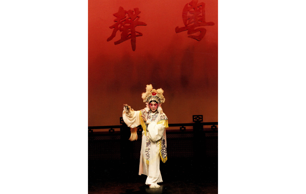 <span style="font-weight: 400;">Un·e artiste portant un costume élaboré blanc et or se tient au centre de la scène devant une toile de fond rouge vif. Deux grands caractères chinois sont projetés sur celle-ci.</span>