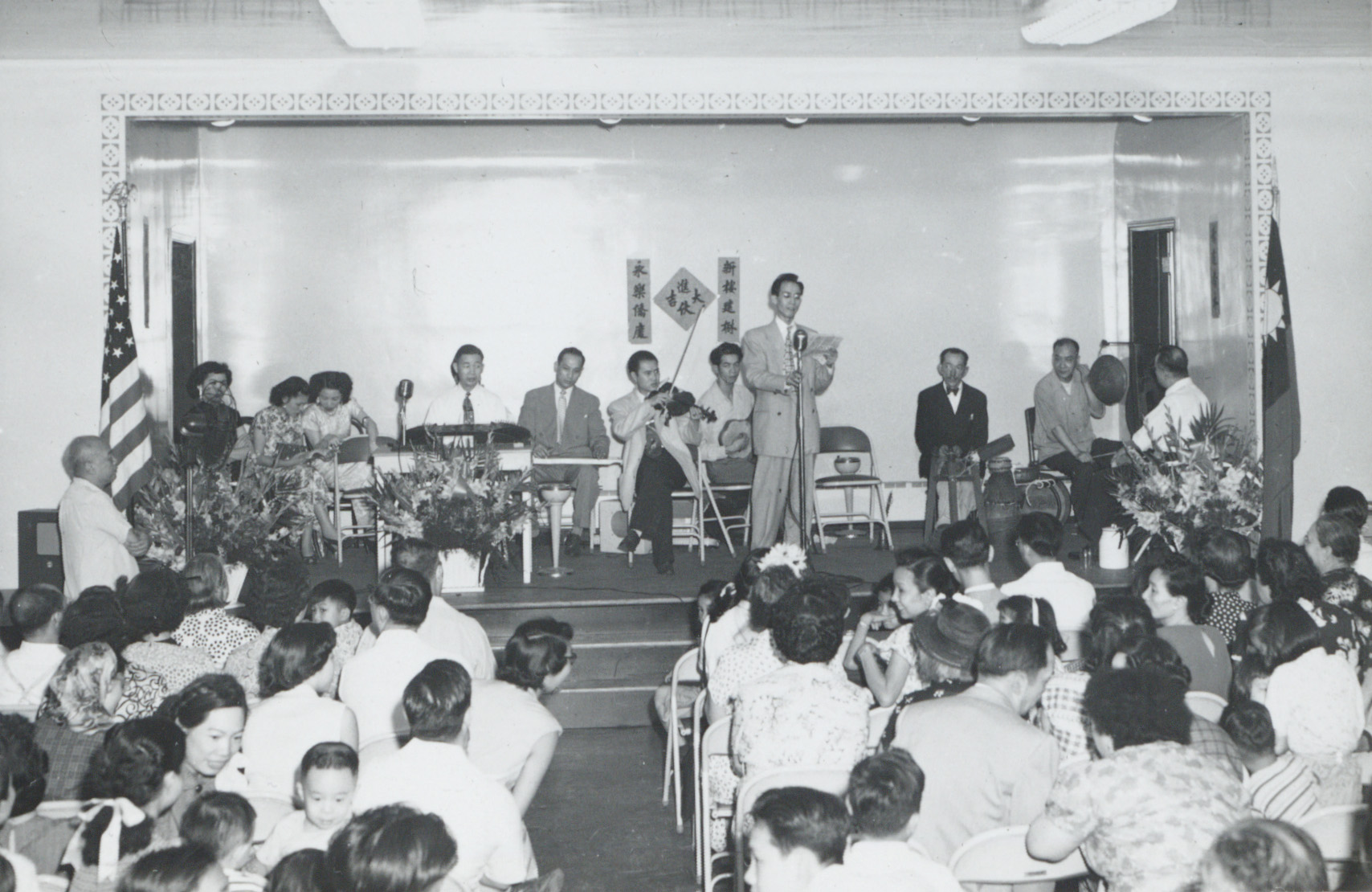<span style="font-weight: 400;">Une salle comble d’hommes, de femmes et d’enfants assiste au concert d’un orchestre cantonais sur une scène sobrement décorée de compositions florales et de petites banderoles.</span>
