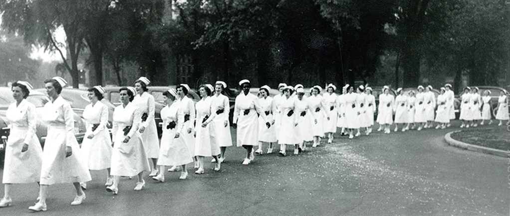 Over 30 women march in a row, wearing white uniforms, caps, and shoes. - Plus de 30 femmes marchent en rangée, portant uniforme, coiffe et souliers blancs.