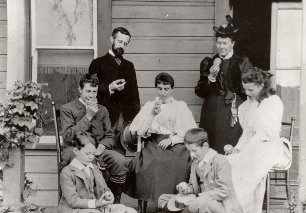 Photo en noir et blanc montrant une famille assise sur une véranda : un homme, deux femmes, une fille et trois garçons qui regardent tous la pomme qu’ils tiennent à la main.