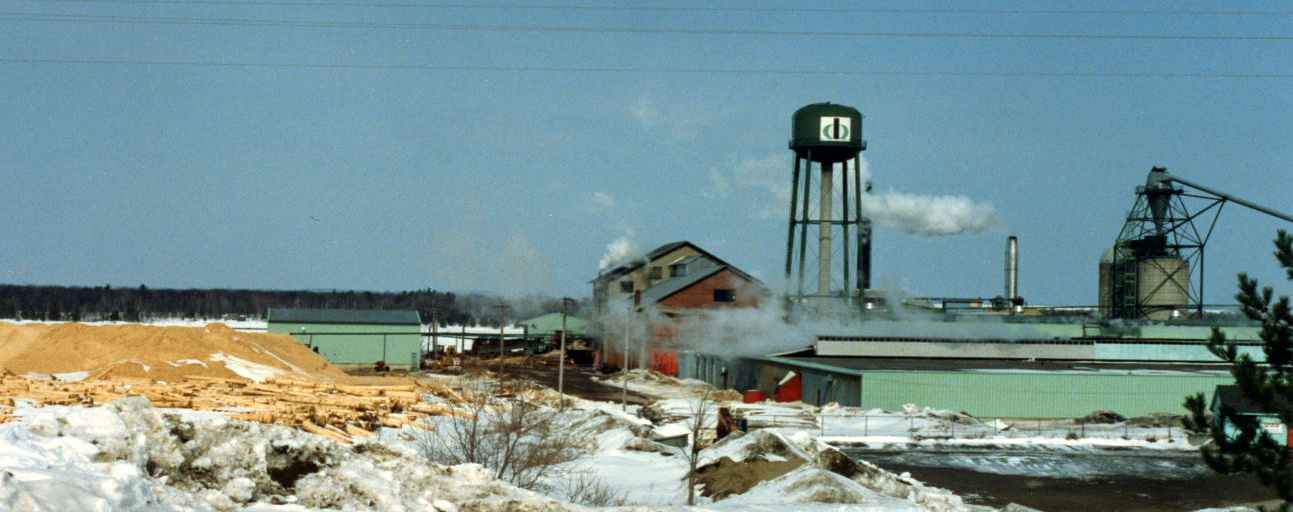 Une scierie faite de briques est entourée de piles de bran de scie l’hiver, avec un château d’eau et un incinérateur à déchets en arrière-plan.