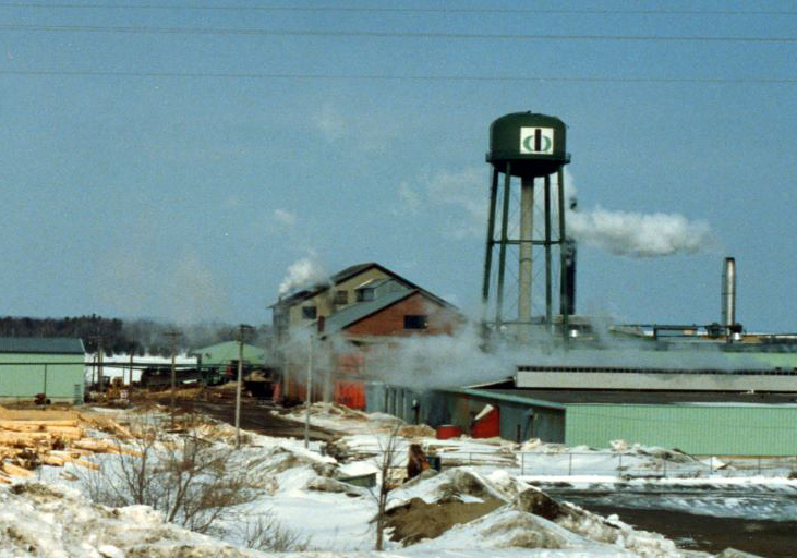 Une scierie faite de briques est entourée de piles de bran de scie l’hiver, avec un château d’eau et un incinérateur à déchets en arrière-plan.