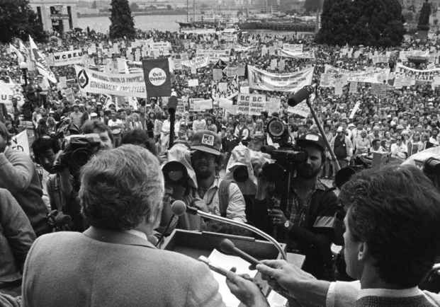 Un homme, prenant la parole devant un podium, fait face à une foule nombreuse sur la pelouse du C.-B. corps législatif. Des journalistes sont rassemblés, tenant des microphones et des caméras vers le podium. Des bannières organisationnelles et des pancartes de protestation sont visibles dans la foule.