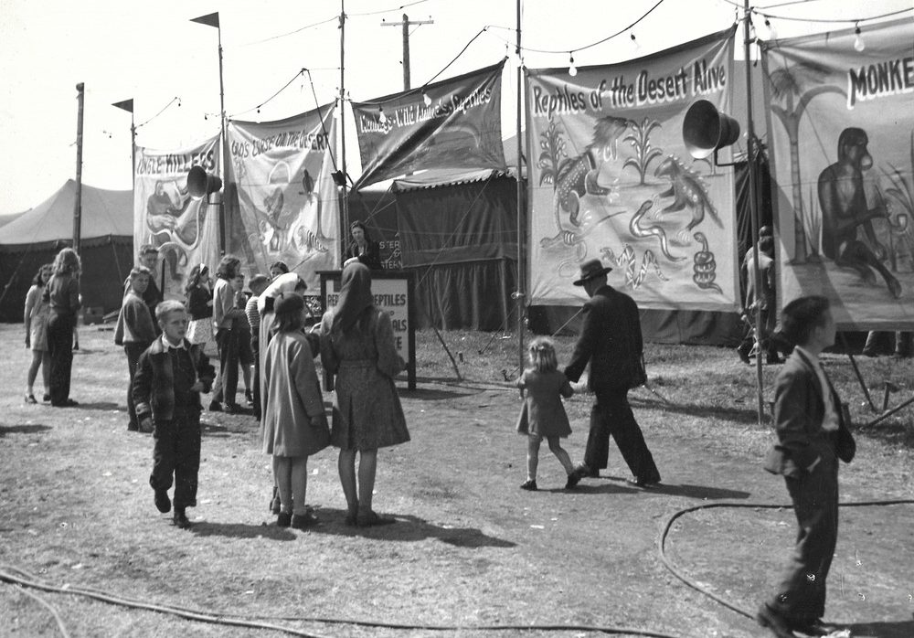 Une photo en noir et blanc d’une foule à une fête foraine parmi de nombreuses tentes; on voit un carrousel en arrière-plan