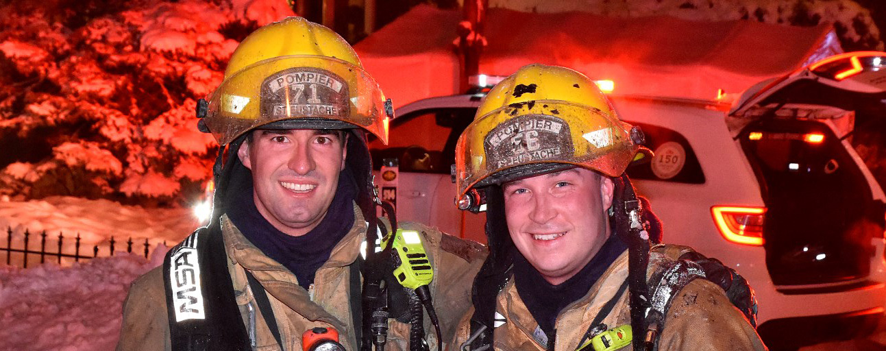 Photographie de deux pompiers souriants en tenue de combat contre le feu, devant un camion de pompiers, une voiture et plusieurs maisons lors d’une nuit éclairée.