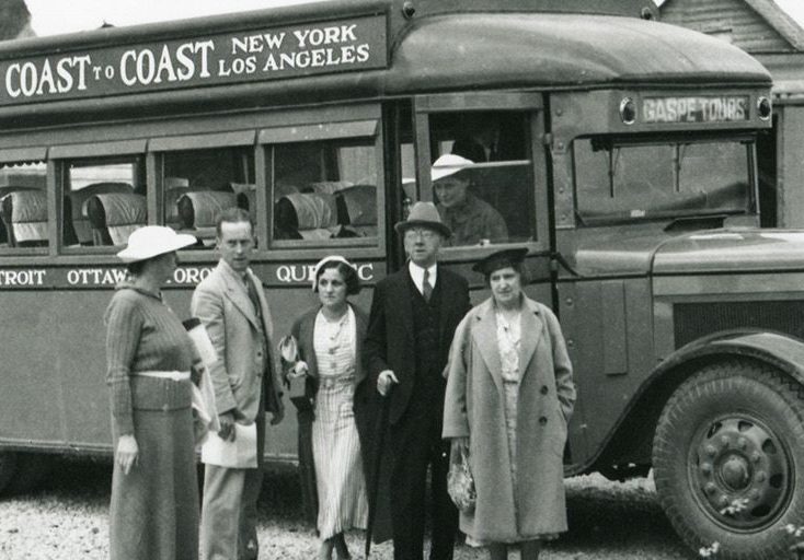 Photographie noir et blanc d’un groupe de personnes devant un autobus voyageur de 1932. Derrière l’autobus, on peut apercevoir quelques maisons du village de Percé et les montagnes qui forment la côte. Sur l’autobus il est écrit : Montreal Vancouver Coast to Coast New York Los Angeles.