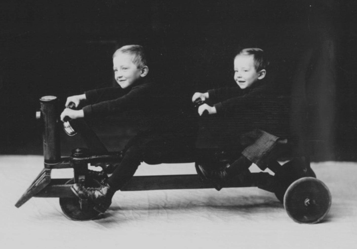 Photo ancienne noir et blanc avec 2 petits enfants souriants assis sur une voiturette à 3 roues avec 2 guidons.