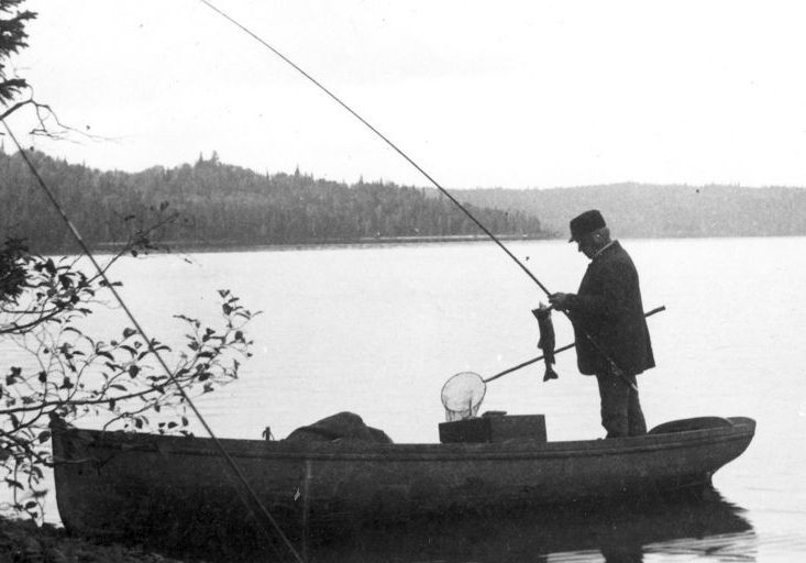 Photo en noir et blanc de pêcheurs dans un bateau à vapeur sur le lac Édouard. Au premier plan, un bateau au bord de la rivière; dedans se tient un homme décrochant un poisson de sa canne.