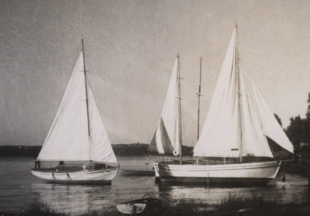 Photographie noir et blanc de trois yachts blancs, aux voiles hissées, ancrés près de la rive