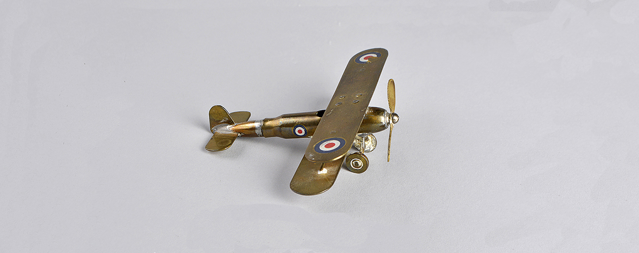 Petit avion en laiton lustré fait avec une cartouche de carabine. L’avion possède une hélice à l’avant et des cocardes sur chaque aile et sur le fuselage.