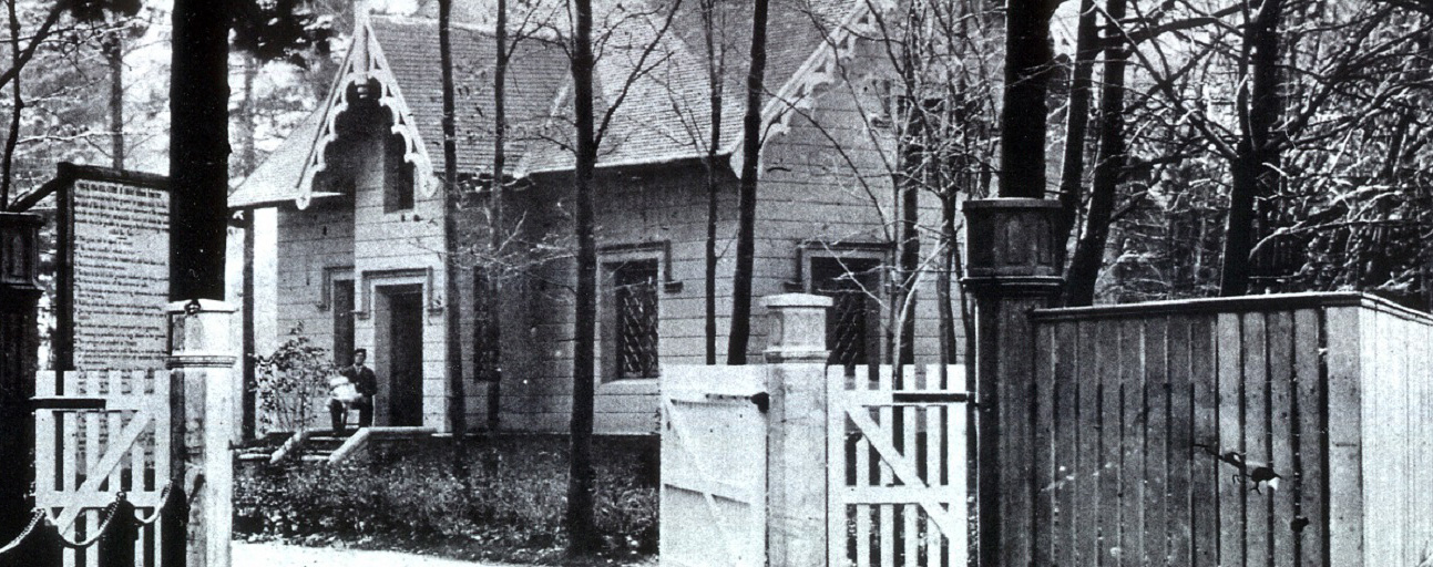 Photo noir et blanc d’une maison entourée d’arbres. Un homme est assis sur le perron tenant un bébé. A l’avant-plan une grande clôture avec une large barrière de bois ouverte.