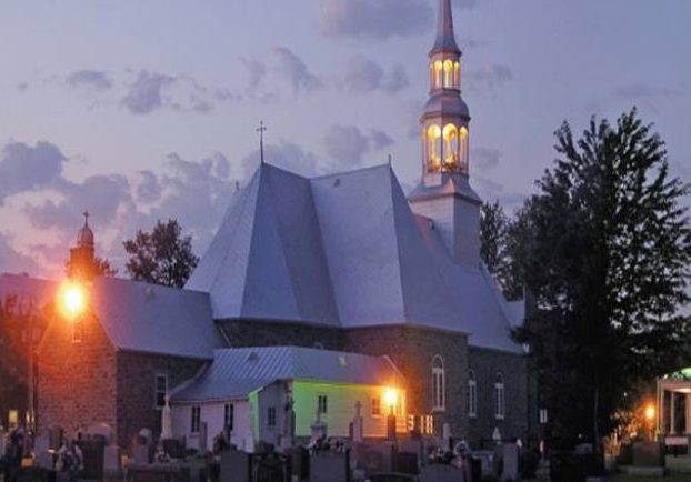 Photographie couleur prise de nuit, plan éloigné de l’arrière des bâtiments comprenant l’église, le presbytère et le cimetière, l’église est éclairée.