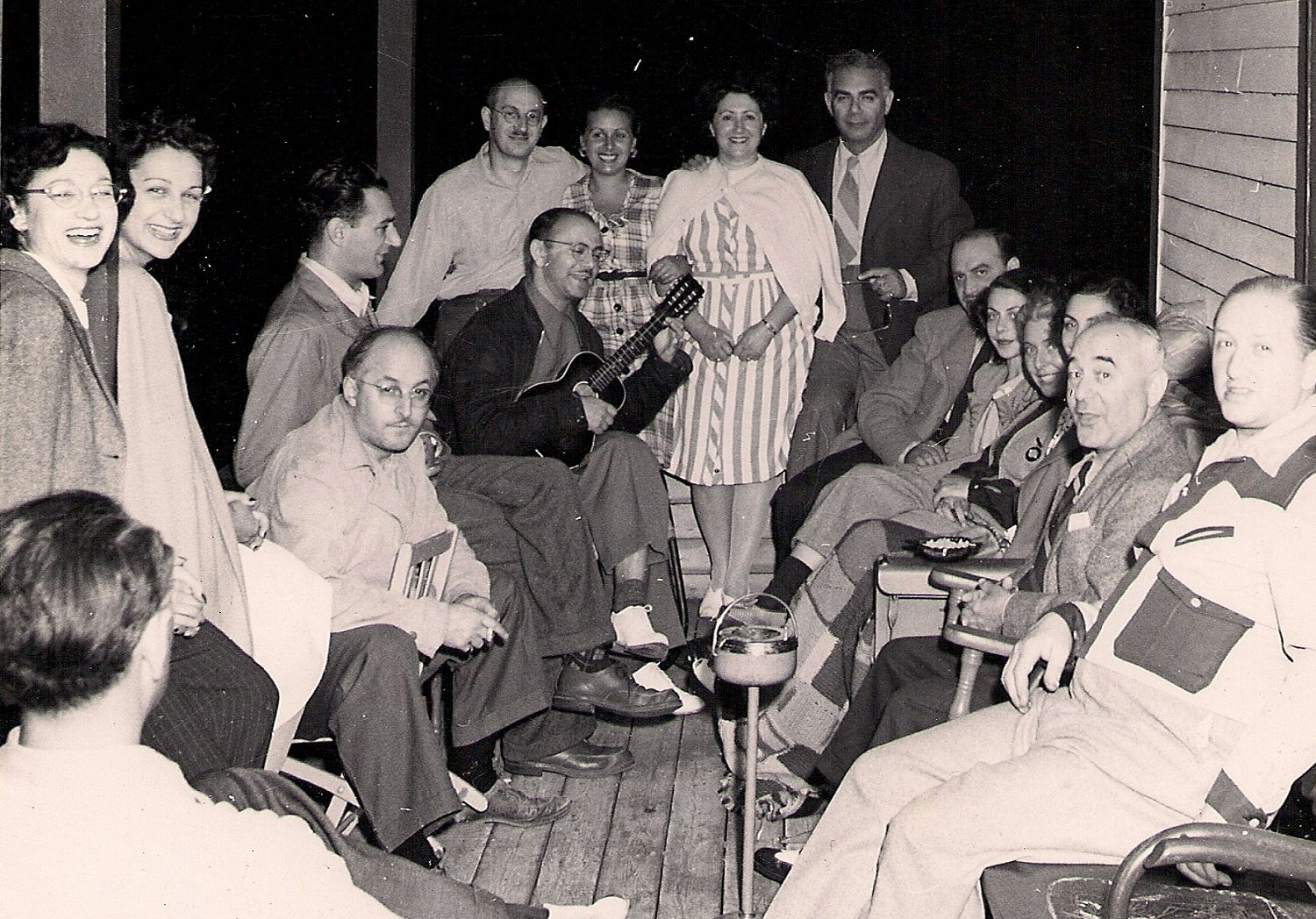 Seize hommes et femmes assis sur une véranda en bois. Un des hommes tient un ukulélé.