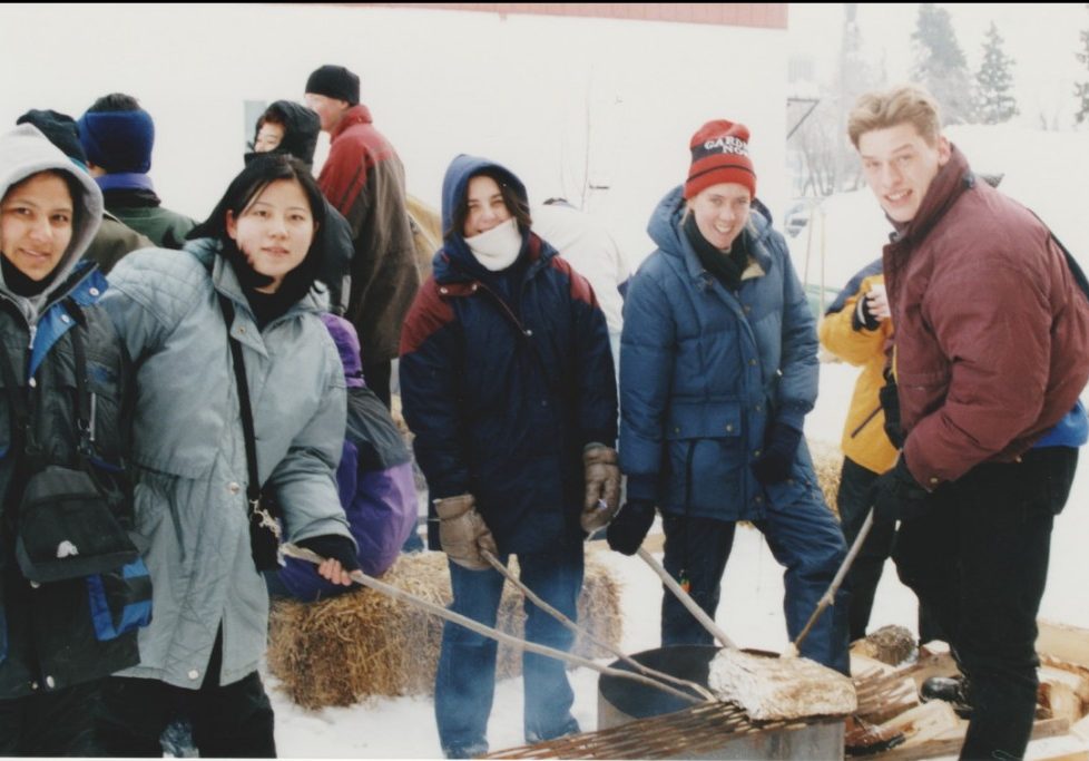 Plusieurs jeunes habillés en vêtements d'hiver qui font griller du pain bannock sur un feu. L'activité faisait partie du festival des trappeurs de 1999.
