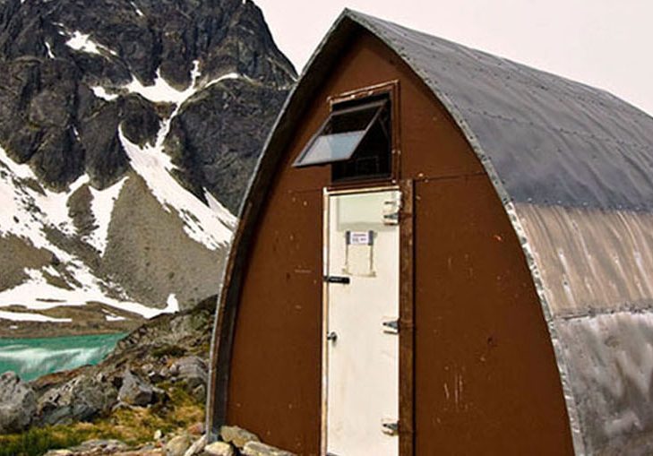 Entrée principale du refuge voûté de style gothique avec sa paroi d’extrémité brune et sa porte blanc brillant, près de l’eau turquoise du lac glaciaire. En arrière-plan, surgit un sommet sombre recouvert de couches de neige éparses.
