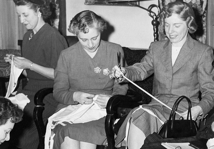 Des femmes travaillent à des œuvres de couture individuelles dans une photo en noir et blanc. Des chaises ont été ajoutées à la pièce pour les accueillir. Une femme est assise à terre dans le coin gauche.