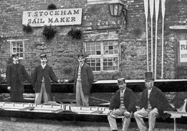 Image des membres de l’équipage de Paris avec leur gérant, J. A. Harding, posant devant un hangar à bateaux à leur arrivée à Southampton, en Angleterre. Cette photographie en noir en blanc datant de juillet 1867 montre un bâtiment fait de blocs de pierre. Leur aviron est devant le bâtiment, les rames appuyées contre la façade.