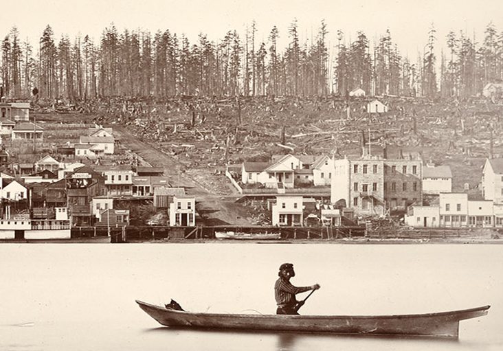 Une photographie monochrome aux tons sépia d’un homme en canot sur le fleuve Fraser passant devant une partie récemment déboisée de New Westminster. New Westminster, à cette époque, compte de nombreux bâtiments près de la rive du fleuve mais, plus on s’éloigne de la rive, moins il y a d’édifices. Le haut des collines est couvert de souches d’arbres.