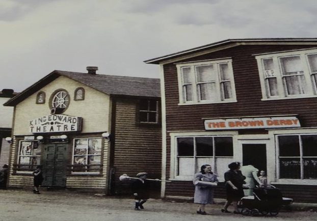 Photographie d'archives couleur. On voit le Brown Derby, le cinéma King Edward, les magasins A. Peckford et S. Cohen & Fils et le café Purity. Kirk Pomeroy porte un balai en bouleau, Daisy Bennett porte un manteau léger et Mme Pomeroy pousse une poussette.