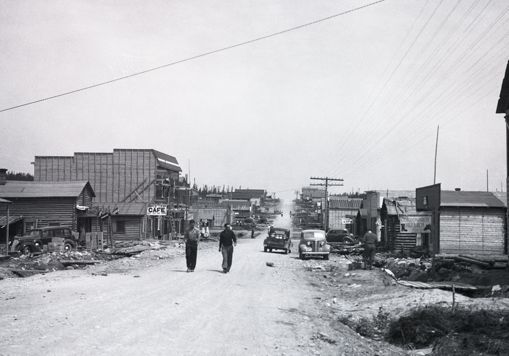 Photographie en noir et blanc d’une route de gravier bordée de bâtiments de planches ou en bois rond. Deux hommes marchent vers la caméra.