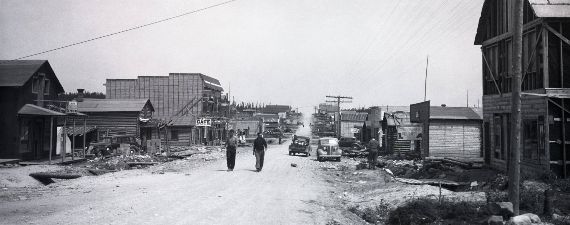 Photographie en noir et blanc d’une route de gravier bordée de bâtiments de planches ou en bois rond. Deux hommes marchent vers la caméra.