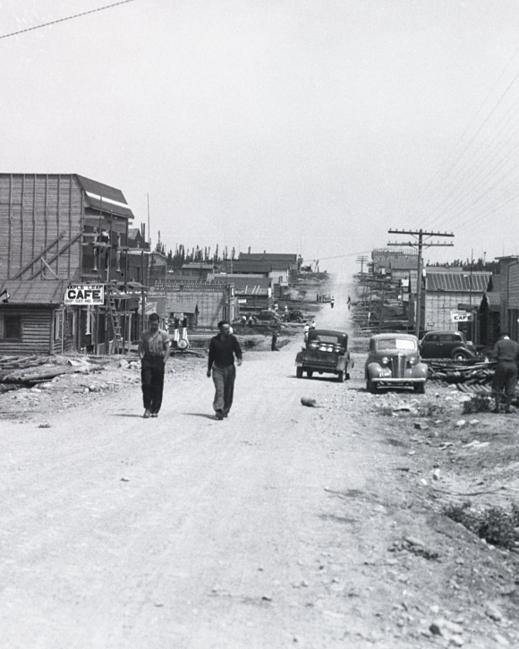 Photographie en noir et blanc d’une route de gravier bordée de bâtiments de planches ou en bois rond. Deux hommes marchent vers la caméra.