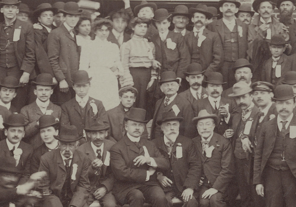 Photographie sépia illustrant des hommes et quelques femmes à l’extérieur devant une usine.