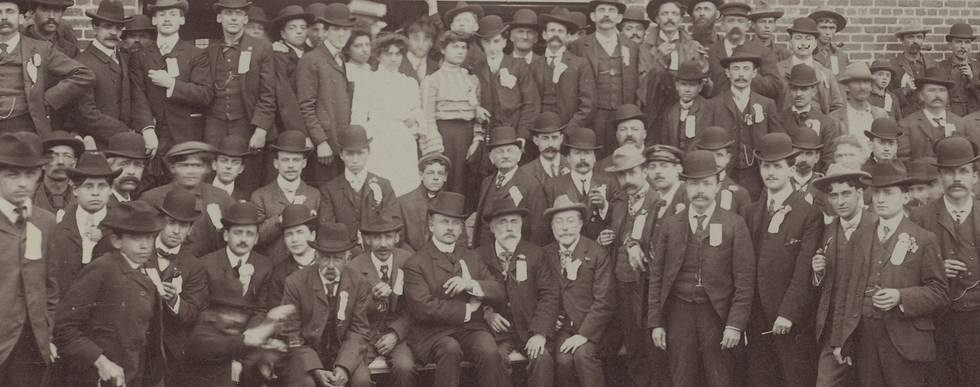 Photographie sépia illustrant des hommes et quelques femmes à l’extérieur devant une usine.