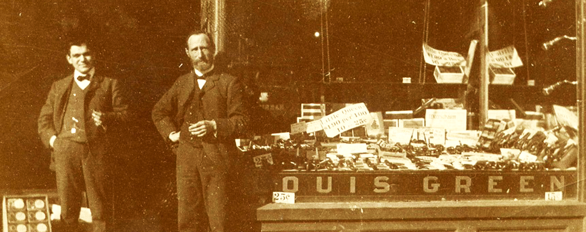 Devanture dotée d’une grande vitrine. En haut, on peut lire « Havana Cigars » et en bas, « Louis Green ». La vitrine expose des cigares et des produits de tabac. Deux hommes se tiennent debout dans l’entrée.