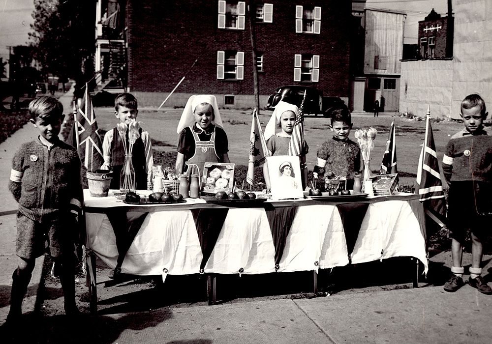 Portrait de six enfants, deux filles au centre et deux garçons de chaque côté, derrière une table remplie de nourriture, quatre drapeaux de l'Union Jack et deux vases de fleurs.
