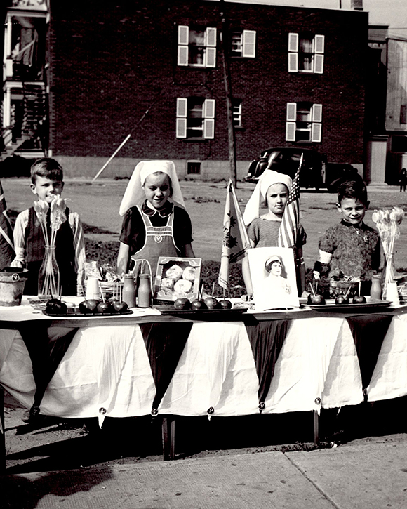 Portrait de quatre enfants, deux filles au centre et un garçon de chaque côté, derrière une table remplie de nourriture, de drapeaux et deux vases de fleurs.