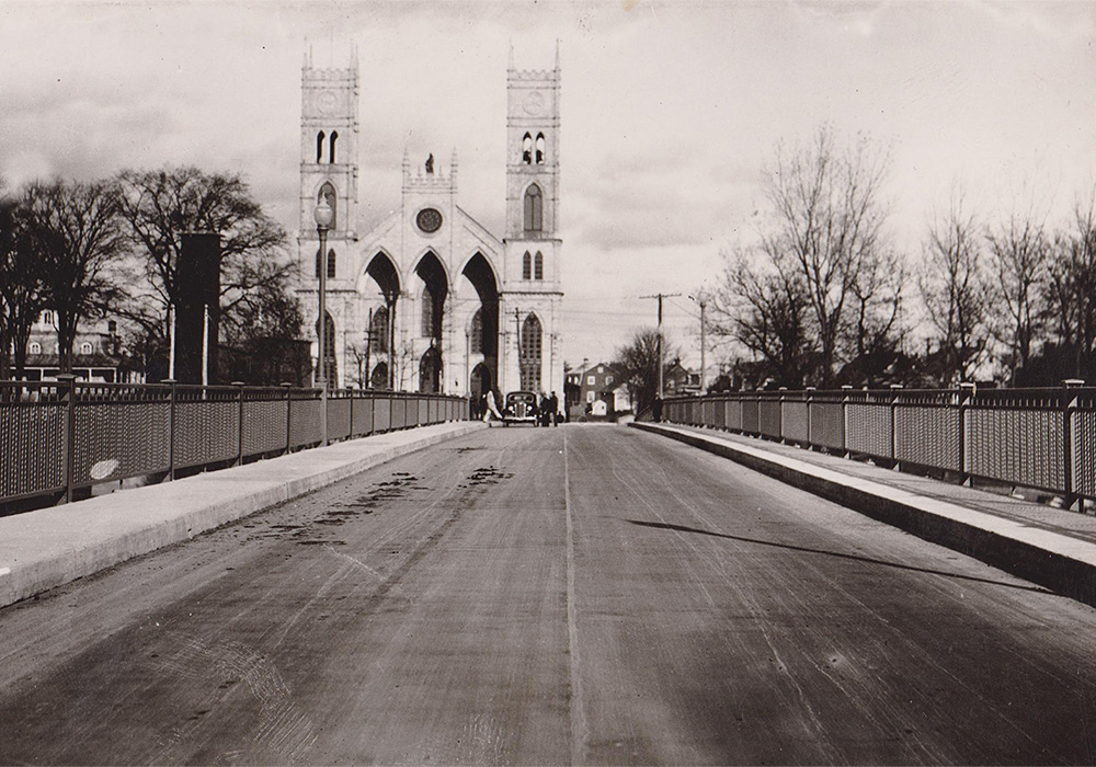 Vue en noir et blanc de la travée d’un pont au bout duquel se dresse une église à deux clochers entourée de maisons anciennes.