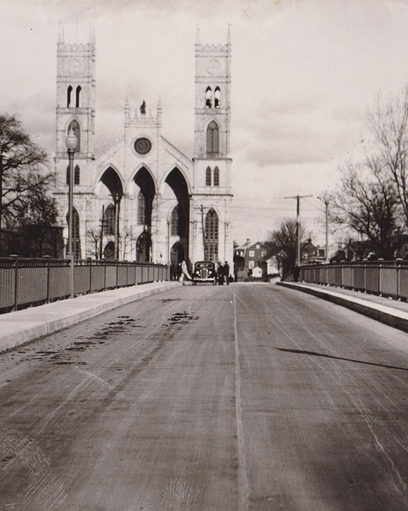 Vue en noir et blanc de la travée d’un pont au bout duquel se dresse une église à deux clochers entourée de maisons anciennes.