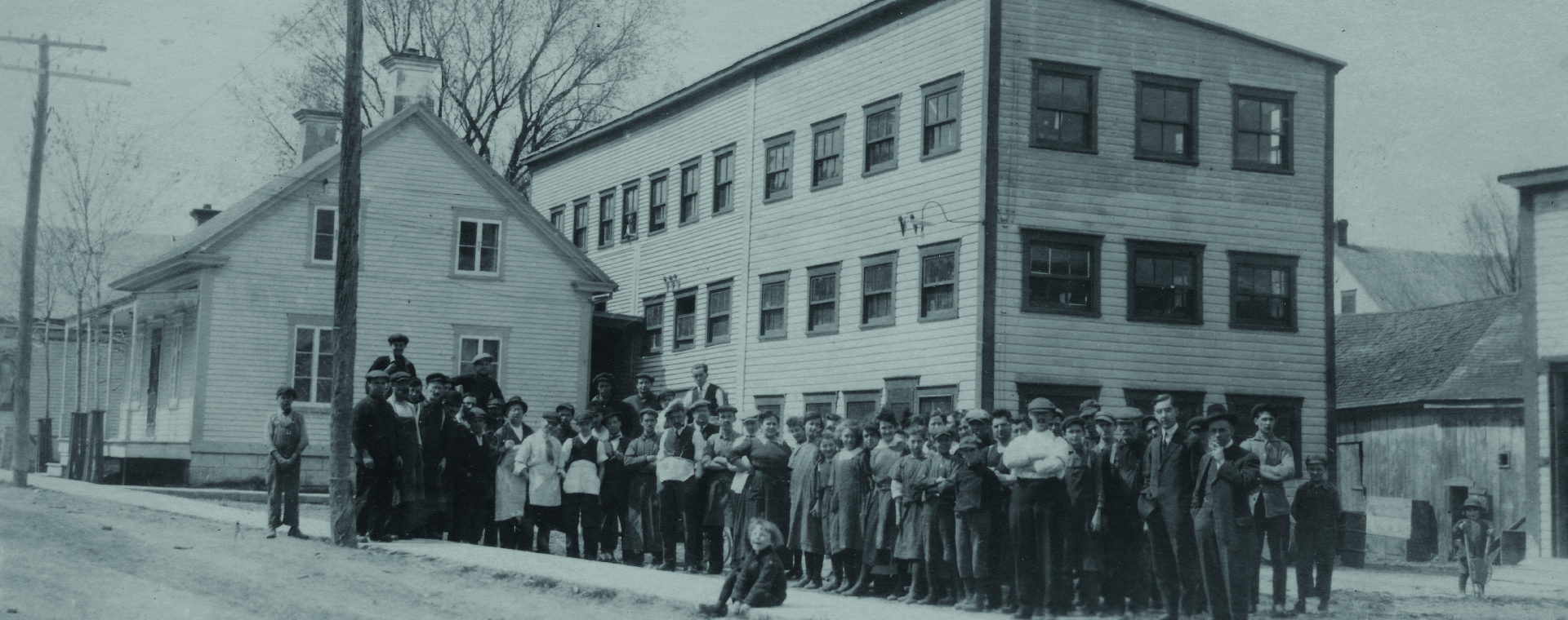 La photo montre les employés rassemblés devant la manufacture Charron. Il s’agit d’une bâtisse en bois de trois étages, construite à l’arrière d’une maison sur la rue St-Antoine.