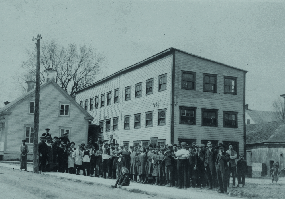 La photo montre les employés rassemblés devant la manufacture Charron. Il s’agit d’une bâtisse en bois de trois étages, construite à l’arrière d’une maison sur la rue St-Antoine.