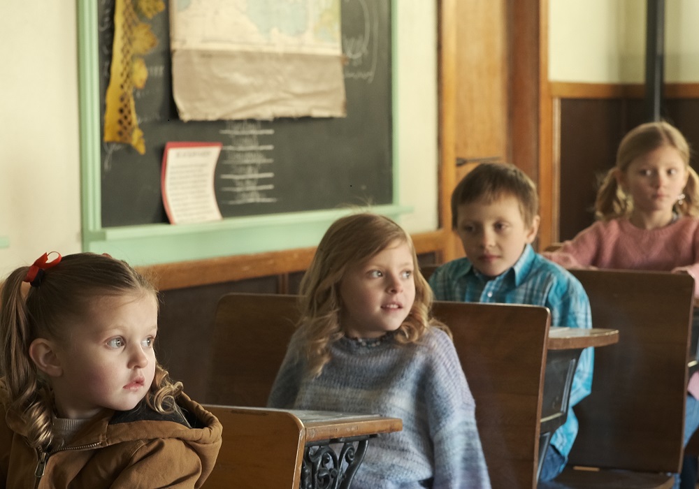 Quatre enfants sont assis sur une rangée de cinq pupitres en bois dans une école de rang.