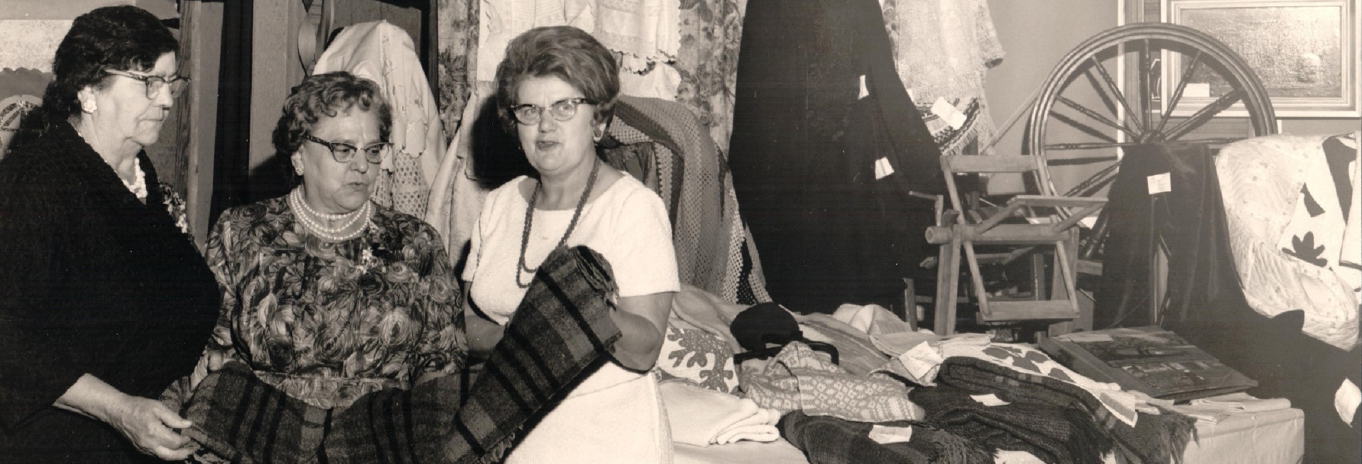 Photographie en noir et blanc de trois femmes examinant une couverture. Derrière elles, une courtepointe et quelques vêtements sont accrochés. À leur droite, des couvertures sont posées sur une table et un rouet est exposé.