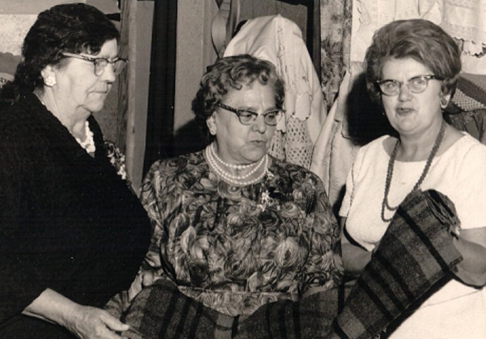 Photographie en noir et blanc de trois femmes examinant une couverture. Derrière elles, une courtepointe et quelques vêtements sont accrochés. À leur droite, des couvertures sont posées sur une table et un rouet est exposé.