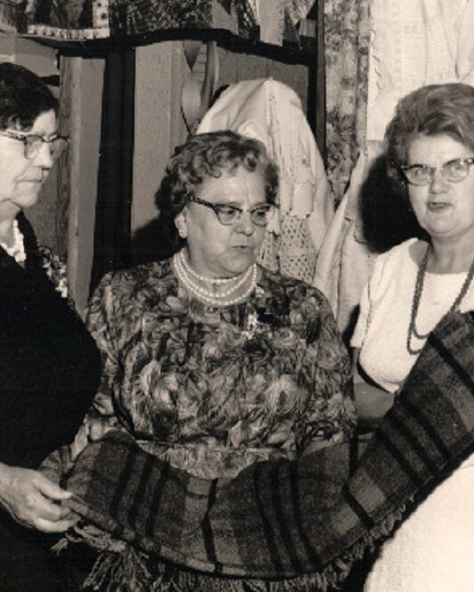 Photographie en noir et blanc de trois femmes examinant une couverture. Derrière elles, une courtepointe et quelques vêtements sont accrochés. À leur droite, des couvertures sont posées sur une table et un rouet est exposé.