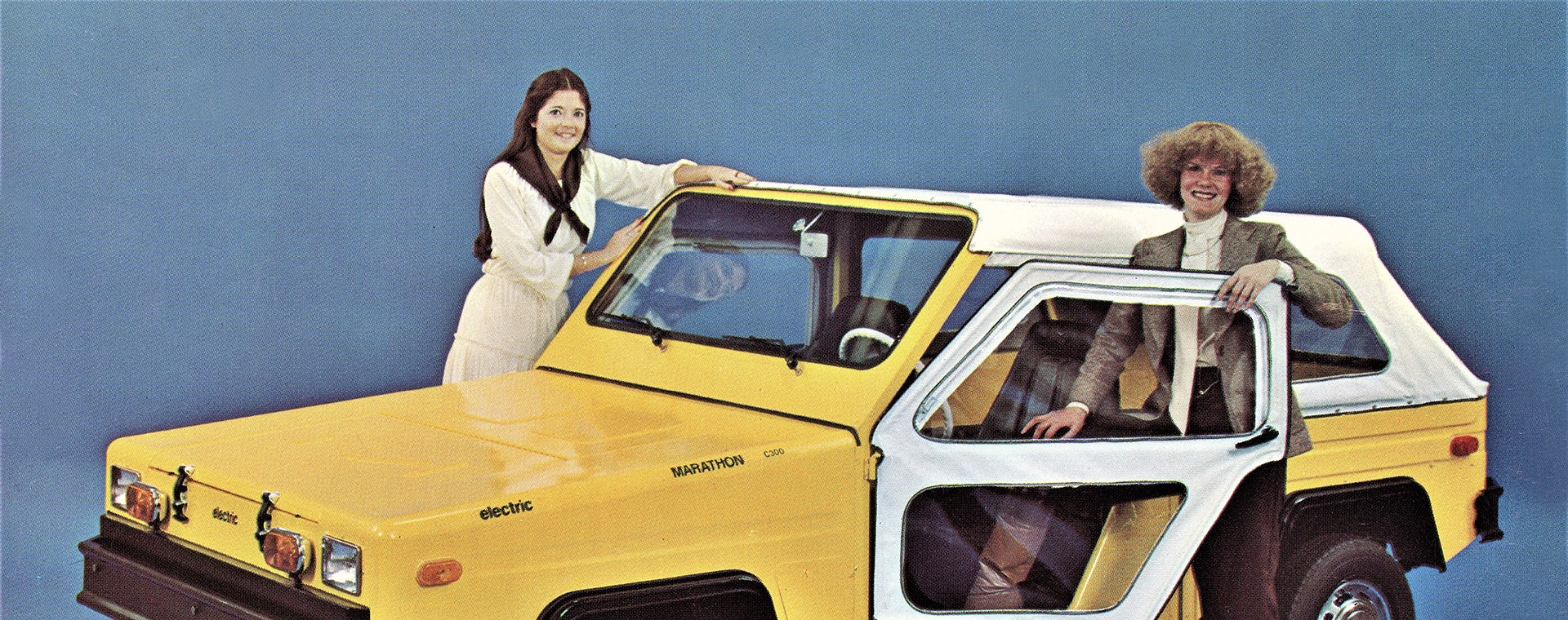 Deux femmes souriantes se tiennent aux portières d'un camion électrique biplace décapotable.