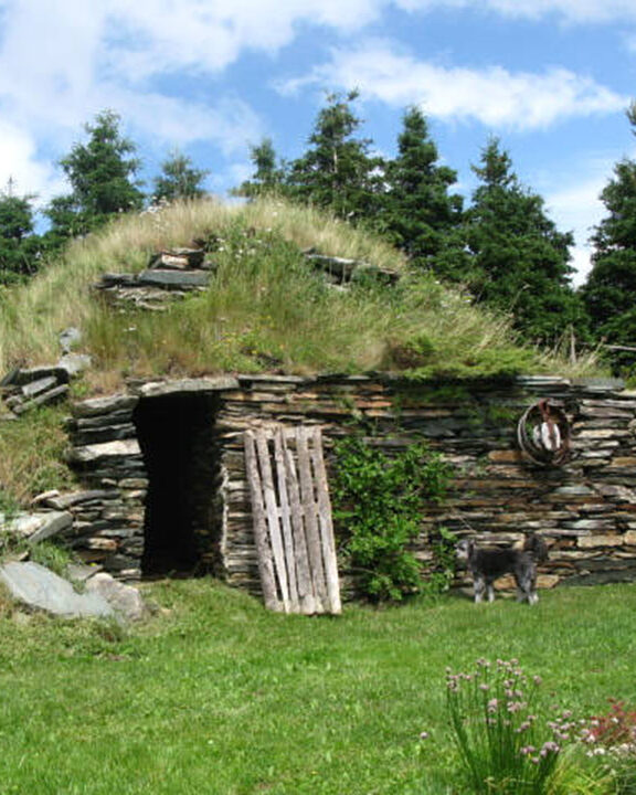 Extérieur d’une cave à légumes à flanc de colline, avec façade en pierres empilées et dessus recouvert d’herbe.